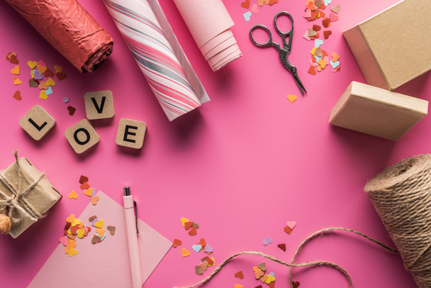 top view of valentines decoration, scissors, gift box, twine, wrapping paper and love lettering on wooden cubes on pink background - Фото, зображення