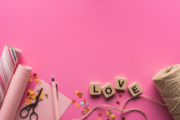 top view of valentines decoration, scissors, wrapping paper, twine and love lettering on wooden cubes on pink background - Photo, Image