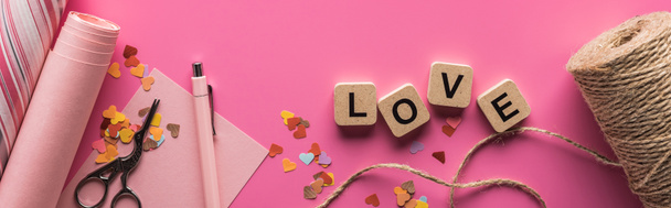 top view of valentines decoration, scissors, wrapping paper, twine and love lettering on wooden cubes on pink background, panoramic shot - Photo, Image