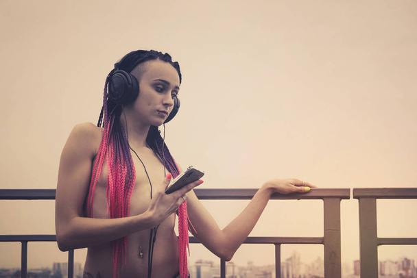 Όμορφη κοπέλα ακούει μουσική με τα ακουστικά της στο δρόμο. Όμορφη άτυπη κοπέλα ντυμένη με σέξι σέξι μαύρο σουτιέν στέκεται κοντά στο κιγκλίδωμα και κοιτάζει την πόλη στο φως το πρωί - Φωτογραφία, εικόνα
