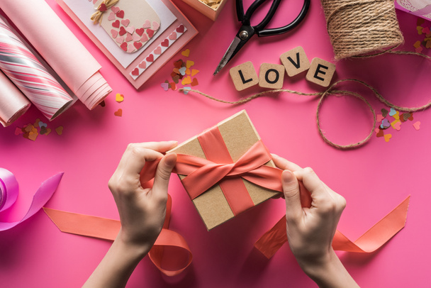 partial view of woman decorating gift box with ribbon near valentines handiwork supplies on pink background - Foto, imagen