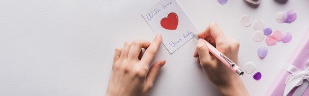 Ausgeschnittene Ansicht einer Frau, die auf Karte neben Valentinstagsgeschenk und Konfetti auf weißem Hintergrund schreibt, Panoramaaufnahme - Foto, Bild