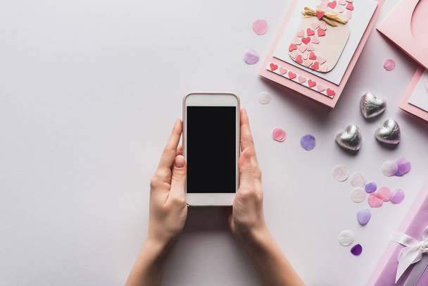 partial view of woman holding smartphone near valentines gifts and decoration on white background - Foto, afbeelding