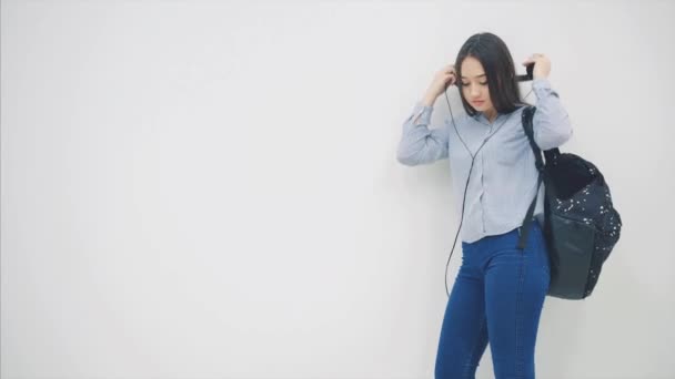 An asian schoolgirl with a backpack appearing on a white background in headphones, taking out and reading notebook. - Séquence, vidéo