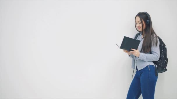 An asian schoolgirl with a backpack appearing on a white background, opening and reading a book, looking happy. - Кадры, видео