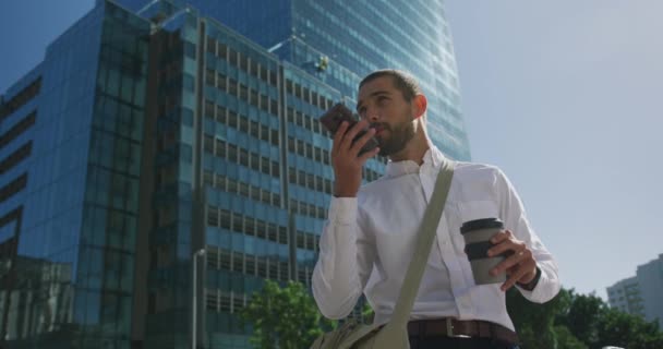Alacsony látószögű kilátás egy fiatal, mosolygó kaukázusi üzletember útközben a városban, beszél az okostelefonon, és kezében egy csésze elvitelre kávé az utcán irodaépület lassított felvétel - Felvétel, videó