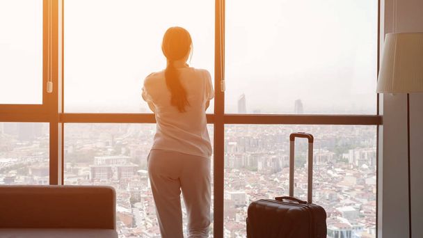 Młoda kobieta podróżniczka z walizką w pokoju hotelowym z panoramicznym widokiem na miasto. - Zdjęcie, obraz