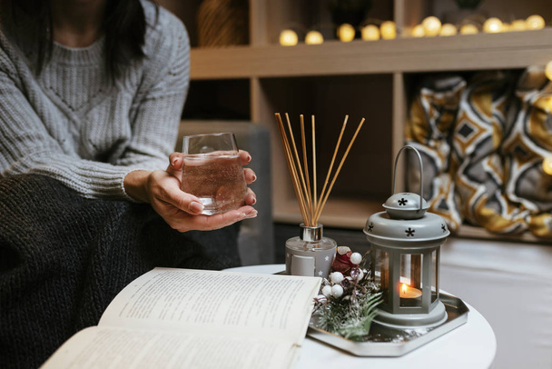 Female in grey knitted sweater holding glass of water wile reading a book - Romantic cozy atmosphere with candles and Christmas lights  - Fotó, kép