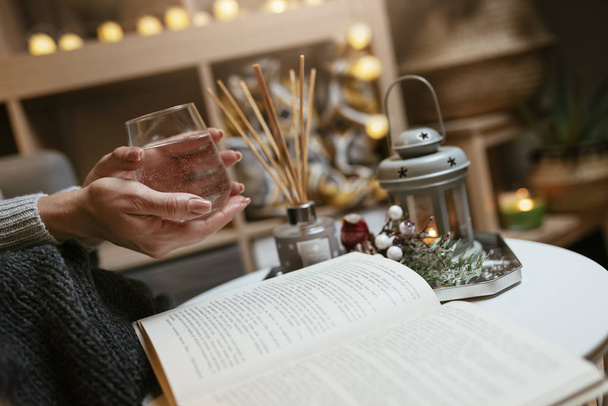 Female in grey knitted sweater holding glass of water wile reading a book - Romantic cozy atmosphere with candles and Christmas lights  - Foto, Imagem