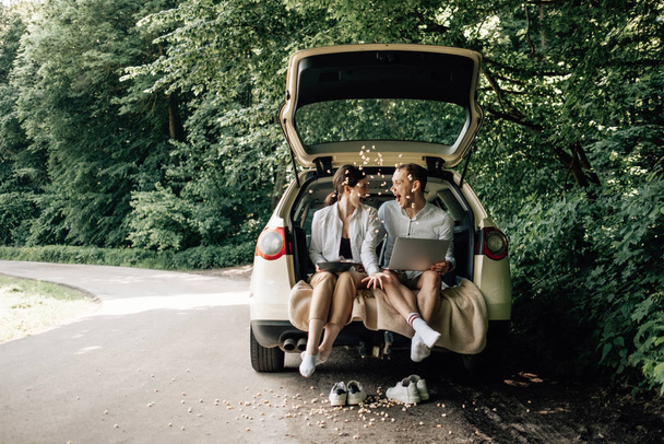 Νεαρό ευτυχισμένο ζευγάρι ντυμένοι Alike σε λευκό T-shirt Καθισμένο στο αυτοκίνητο Trunk με Laptop και Popcorn στην άκρη του δρόμου, Σαββατοκύριακο έξω από την πόλη, Διακοπές και Road Trip Concept - Φωτογραφία, εικόνα