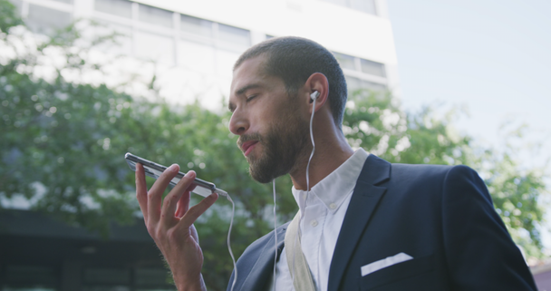 Low angle side view of a young Caucasian businessman on the go in the city, with earphones on talking on the smartphone with office building in the background, slow motion - Materiaali, video