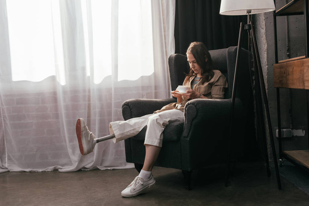 自宅でアームチェアでコーヒーを飲む脚の義肢を持つ若い女性 - 写真・画像