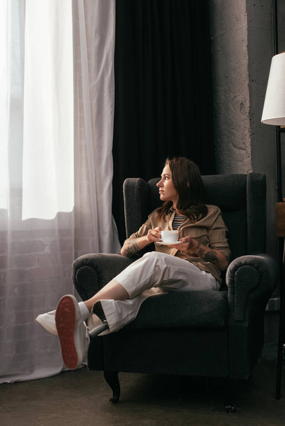Chica con prótesis de pierna sosteniendo taza de café y mirando a la ventana en la sala de estar
 - Foto, imagen