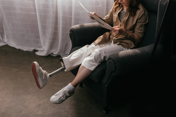 Cropped view of young woman with leg prosthesis reading newspaper in armchair at home - Photo, Image