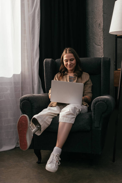 Mosolygó fiatal nő műlábú segítségével laptop a nappaliban - Fotó, kép