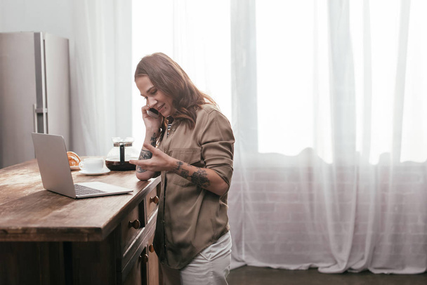 Vista lateral de la mujer sonriente hablando en el teléfono inteligente junto a la computadora portátil en la mesa de la cocina
 - Foto, imagen