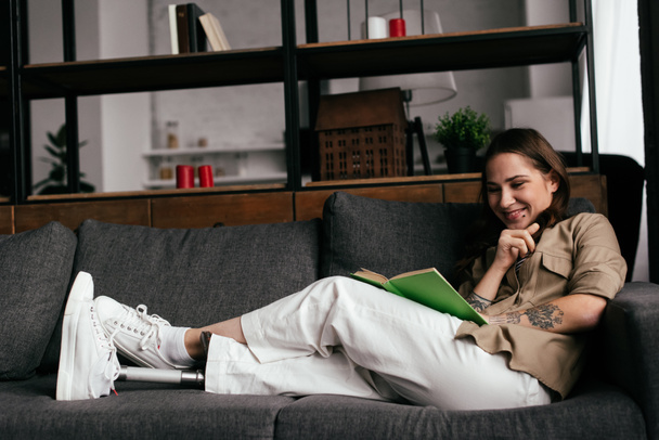 Mujer sonriente con prótesis de pierna libro de lectura en el sofá en la sala de estar
 - Foto, Imagen