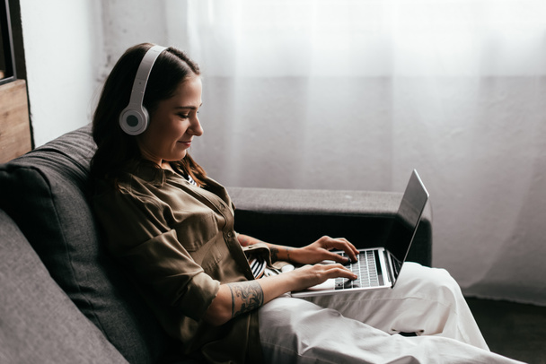 Oldalnézet mosolygós szabadúszó a fejhallgató dolgozik laptop kanapén - Fotó, kép