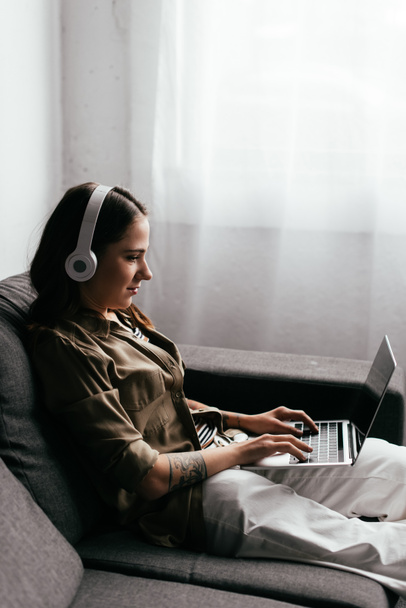 Side view of woman in headphones using laptop on sofa - Photo, Image