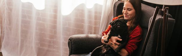 Sonriente chica sosteniendo perro pug en sillón en casa, tiro panorámico
 - Foto, Imagen