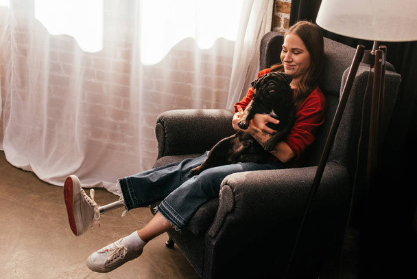 Smiling woman with prosthetic leg holding pug dog in armchair at home, panoramic shot - Foto, afbeelding