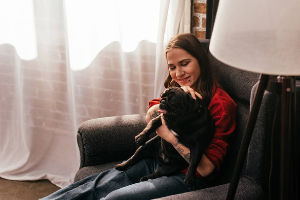 Vrolijke vrouw met kunstbeen aaien pug hond in fauteuil thuis - Foto, afbeelding