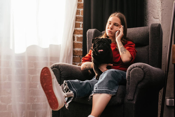 Girl view prosthetic leg talking on smartphone and holding pug in armchair - Foto, Imagen