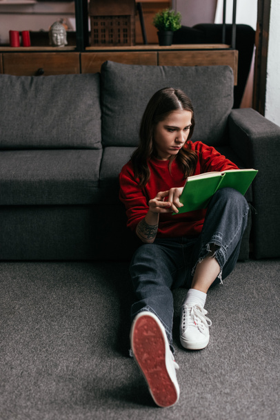 Приваблива молода жінка читає книгу на підлозі біля дивана вдома
 - Фото, зображення