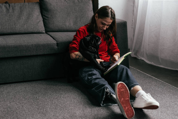 Kobieta z protezą nogi czytania książki i trzymając mopsa na podłodze w salonie - Zdjęcie, obraz