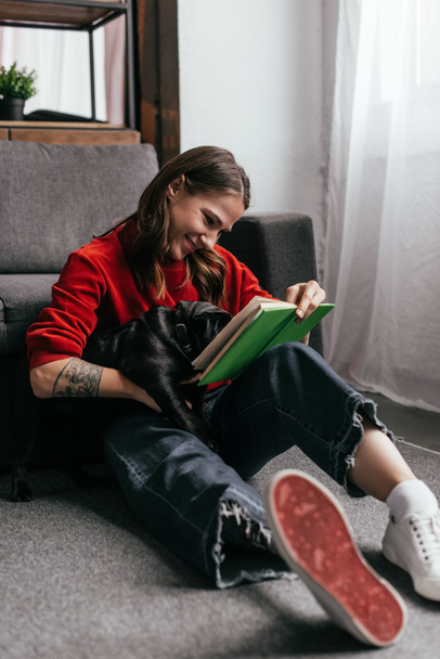 Улыбающаяся девушка с протезной ногой читает книгу и обнимает мопса на полу дома
 - Фото, изображение