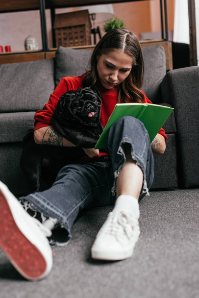 Enfoque selectivo de chica atractiva con pierna protésica abrazando pug y lectura de libro en el suelo en casa
 - Foto, Imagen