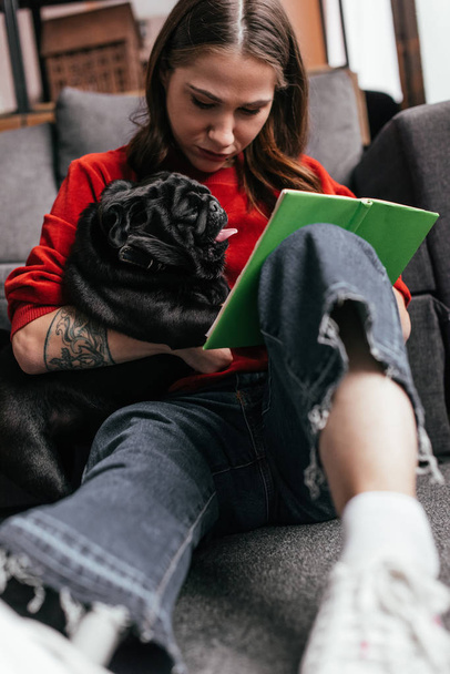 Selective focus of girl with leg prosthesis sitting on floor with pug and book - Fotó, kép