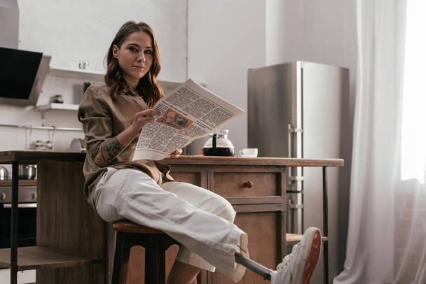 Młoda kobieta z protezą nogi trzyma gazetę i patrzy w kamerę przy stole kuchennym - Zdjęcie, obraz