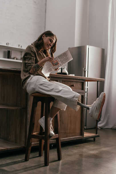 Mujer joven sonriente con prótesis de pierna leyendo periódico en la cocina
 - Foto, imagen