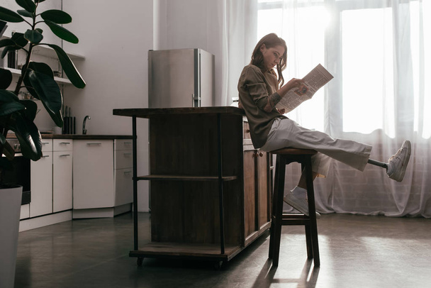 Вид сбоку женщины с протезом ноги, читающей газету за кухонным столом
 - Фото, изображение