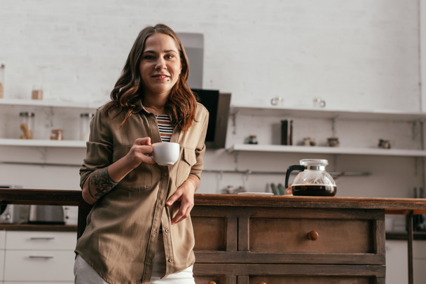 Beautiful girl holding coffee cup and smiling at camera on kitchen - Foto, Imagem