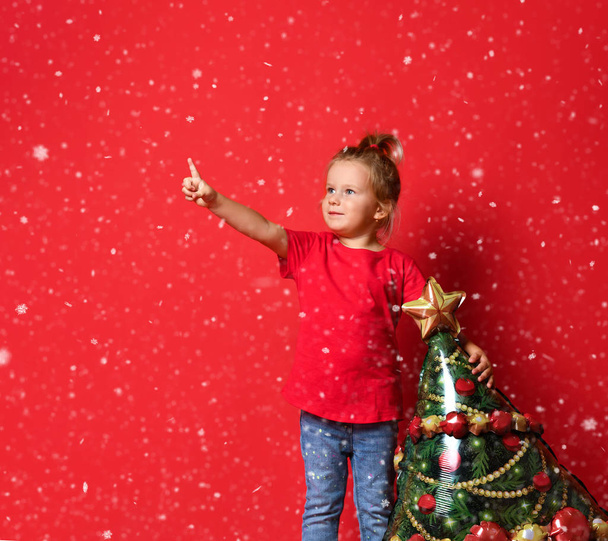 Happy toddler girl in a red T-shirt, blue jeans holding a Christmas tree balloon on a red background with free copy space. - Fotoğraf, Görsel