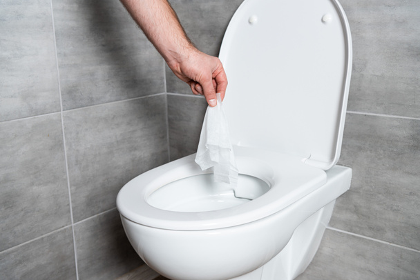 Oříznutý pohled na muže házet ubrousek v záchodové míse v koupelně s šedou dlaždice - Fotografie, Obrázek