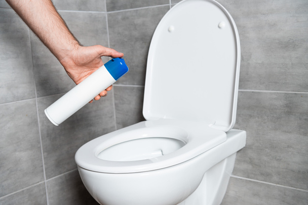 Vista cortada do homem segurando ambientador acima vaso sanitário no banheiro com azulejo cinza
 - Foto, Imagem