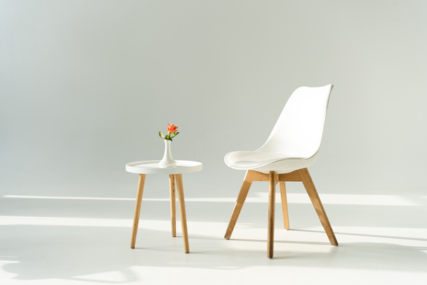 White trendy chair by table with rose in vase on grey background - Foto, Imagen