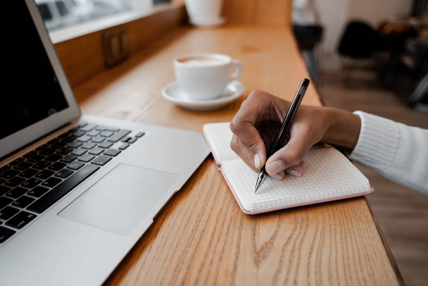 Письменник робить нотатки в блокноті з ручкою, використовуючи ноутбук в місцевому кафе і пити каву. Концепція цифрових технологій та робота фрілансера віддалено. Концепція способу життя людей
 - Фото, зображення