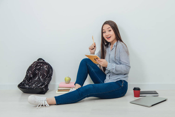 Cute young asian girl is sitting on the floor, holding notebook on her knee, having a nice idea what to write. A lot of school supplies around her. - Photo, image