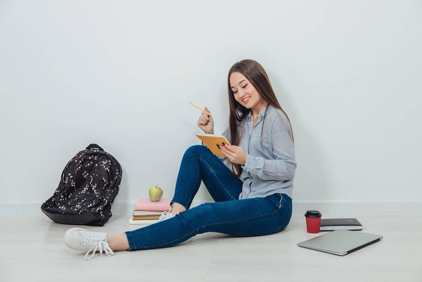 Lovely smiling asian student girl sitting on the floor in lotos position, holding an apple. A lot of school supplies around her. - Photo, image