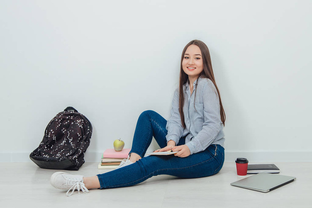 Cute young asian girl is sitting on the floor, holding notebook on her knee, looking lovely at the camera. A lot of school supplies around her. - Foto, afbeelding