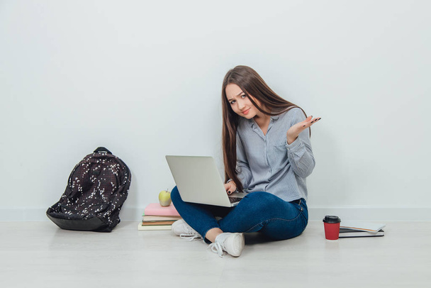 Lovely young asian lady sitting on the floor in lotos position, searching something in her laptop. Puzzled face expression. School supplies around her. - Foto, Imagen