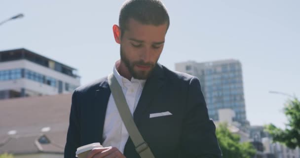 Front view of a young Caucasian businessman on the go in the city, checking time on smartwatch and holding a cup of takeaway coffee with buildings behind him, slow motion - Filmagem, Vídeo