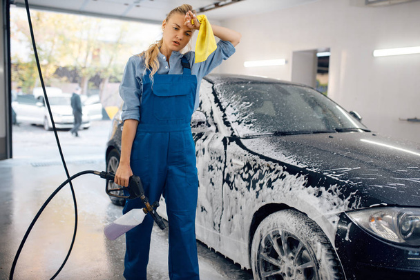 Tired female washer in uniform with foam gun in hands, car wash. Woman washes vehicle, carwash station, car-wash business - Photo, Image