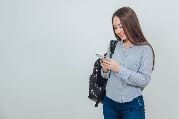 Young lovely asian student standing, holding a phone, smiling, messaging with someone, backpack slung over her shoulder. - Φωτογραφία, εικόνα
