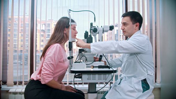 augenärztliche Behandlung - junge hübsche lächelnde Frau überprüft ihre Sehschärfe mit einem Spezialgerät im geräumigen Schrank - legt ihr Kinn auf den Ständer - Foto, Bild