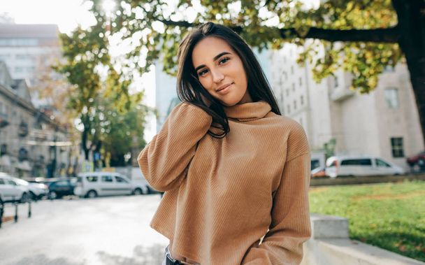 Outdoor image of beautiful young brunette woman wearing beige sweatshirt, smiling and looking directly to the camera. Pretty female student posing in the city street.  - Foto, imagen